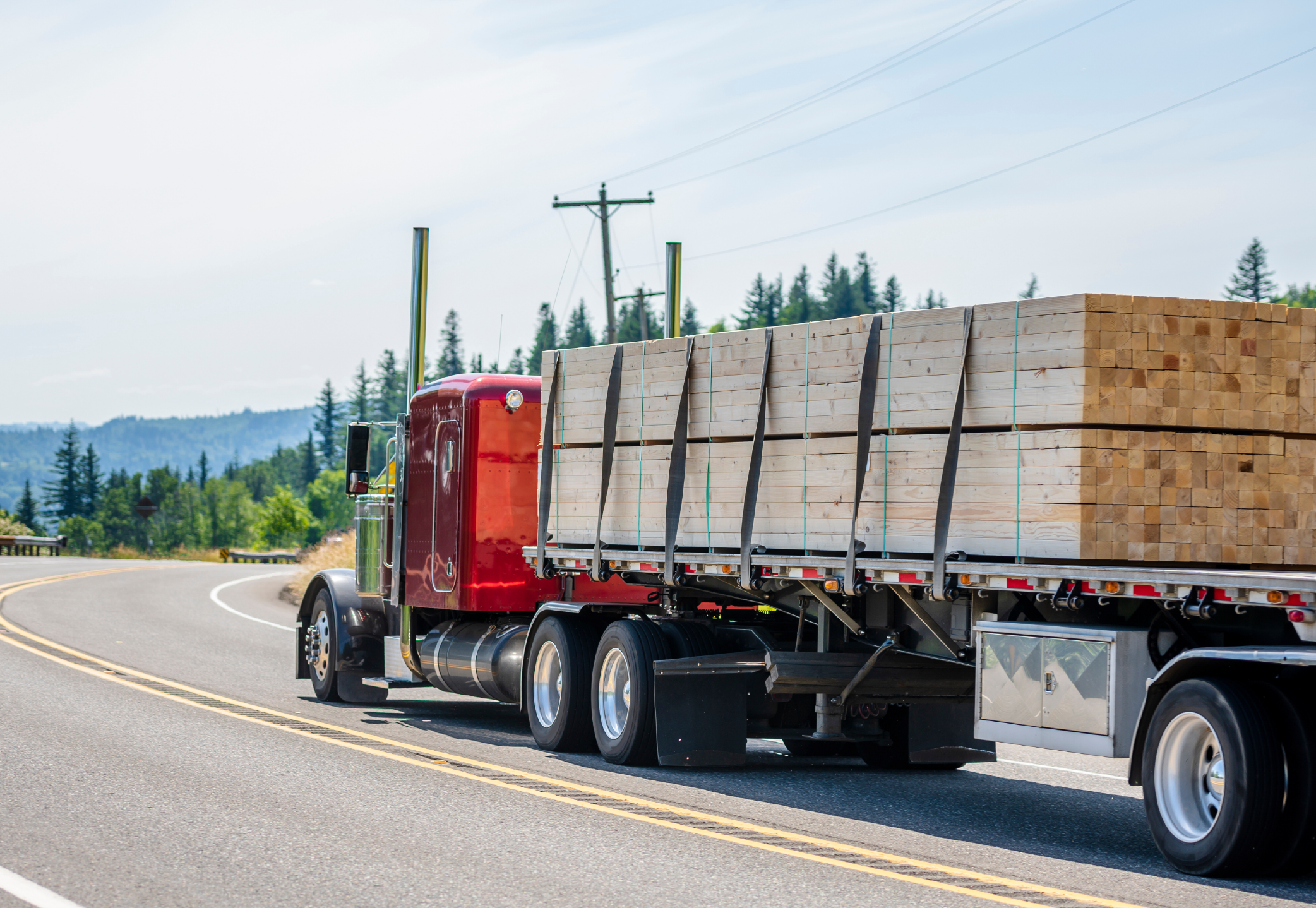 Forestry & Building LP_Image 2 (1) A red semi-truck with a flatbed trailer hauls a load of neatly stacked wooden beams secured with black straps. The truck is traveling on a two-lane rural road with a scenic backdrop of green trees and hills. Utility poles and a guardrail line the road, highlighting transportation and logistics in the lumber or construction industry.